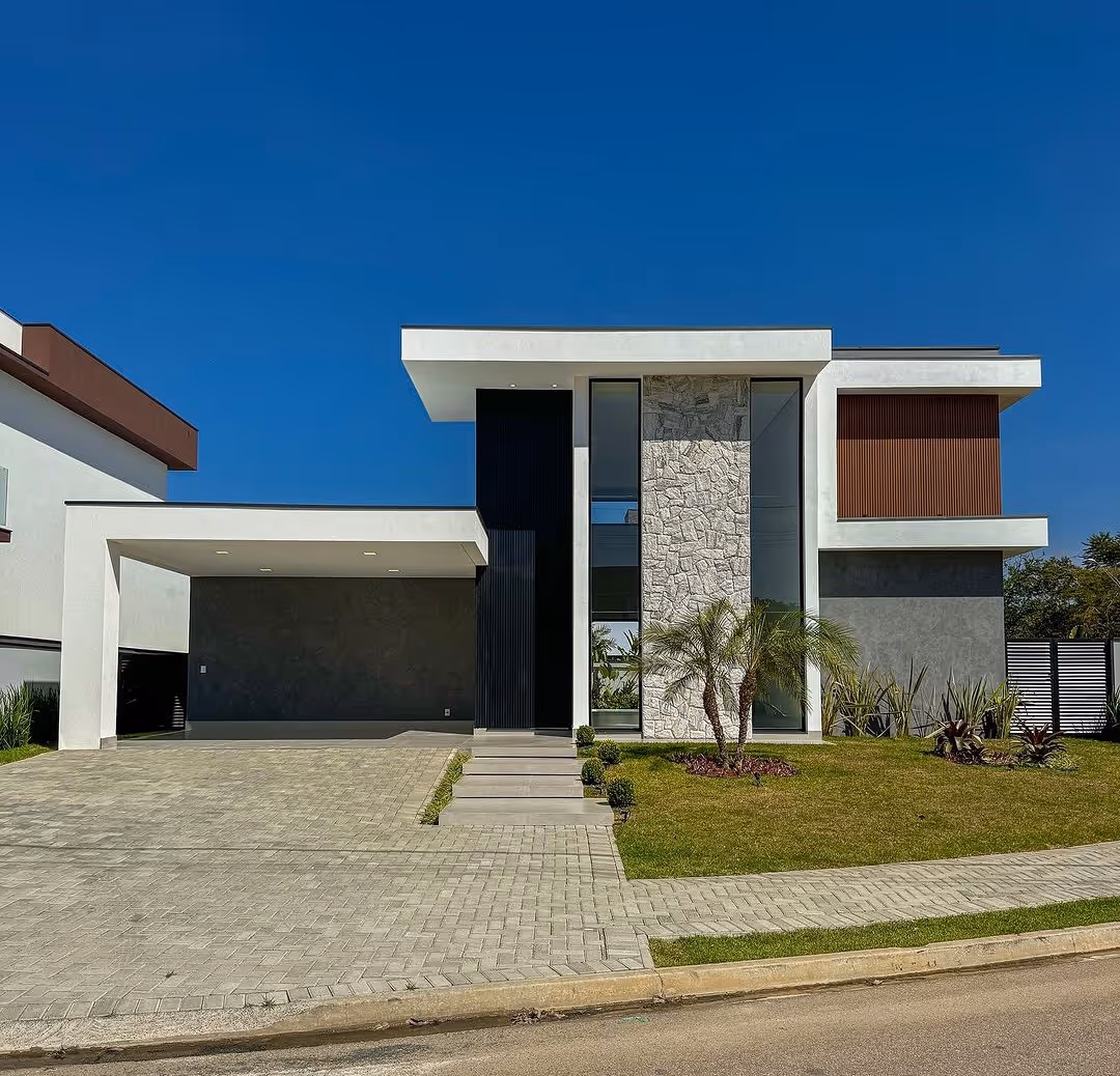 Casa moderna de dois andares com fachada em tons neutros, jardim com palmeiras e garagem ampla sob céu azul.