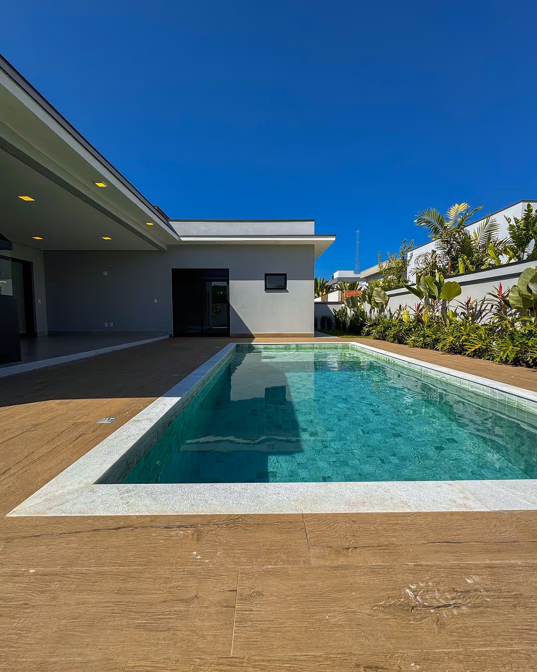 Piscina retangular de água azul clara cercada por piso de madeira com casa moderna e jardim ao fundo sob céu azul.