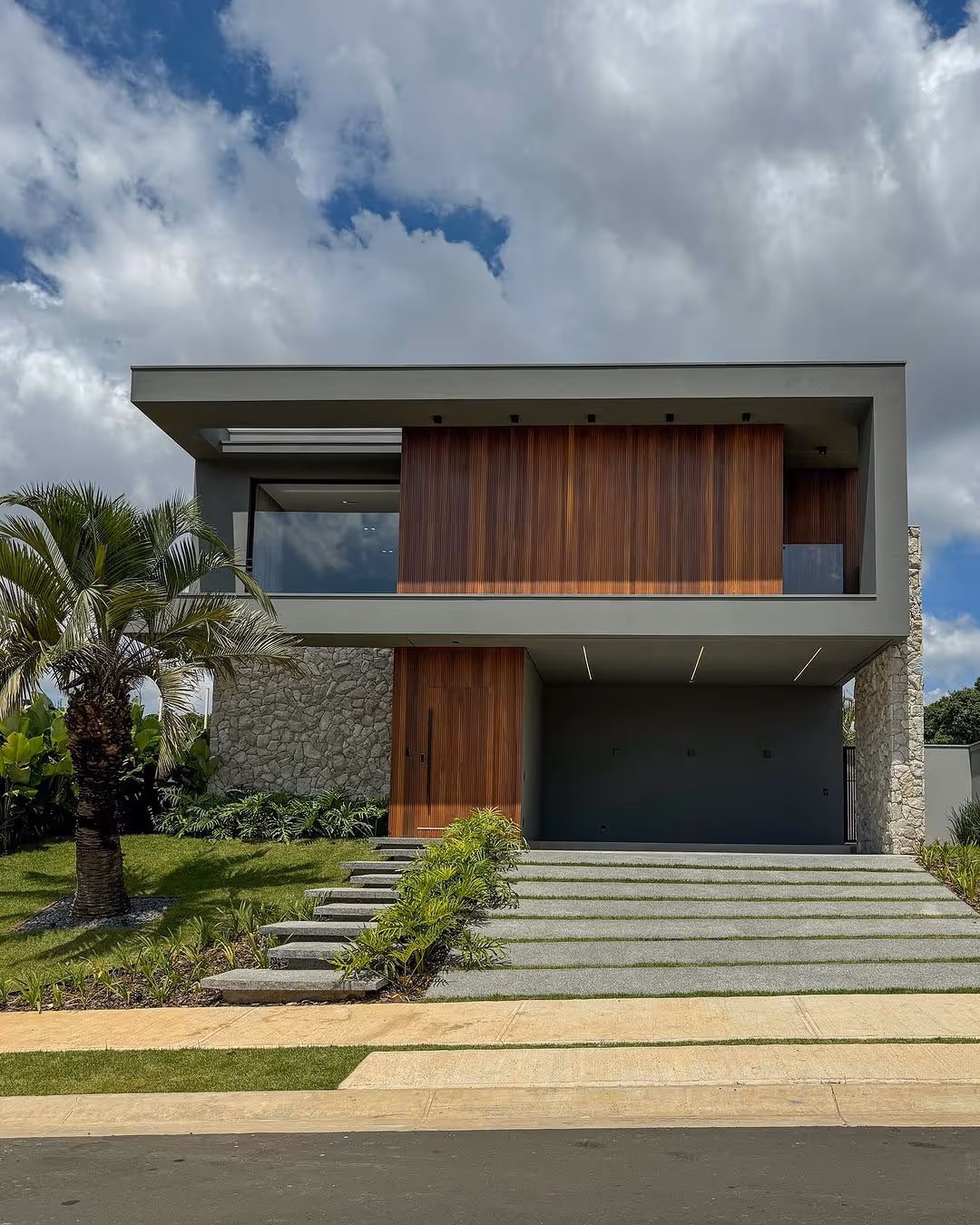 Casa moderna de concreto com grandes janelas e plantas penduradas, cercada por árvores e vegetação seca.