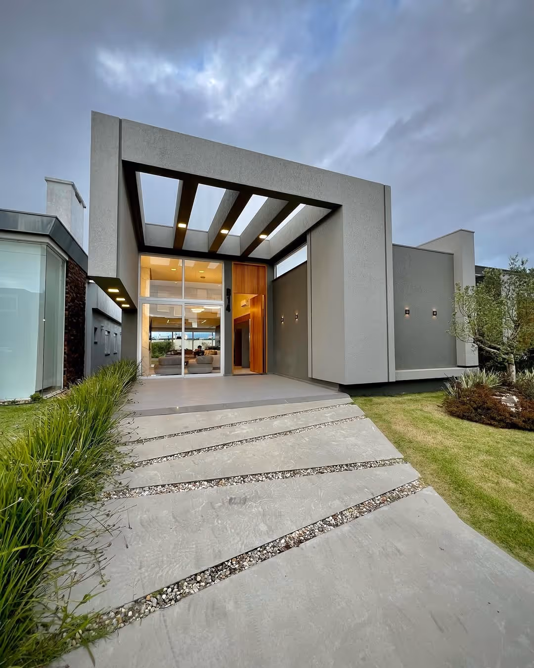 Fachada moderna de casa cinza com laje aberta, iluminação embutida, vidro na entrada e jardim ao redor.