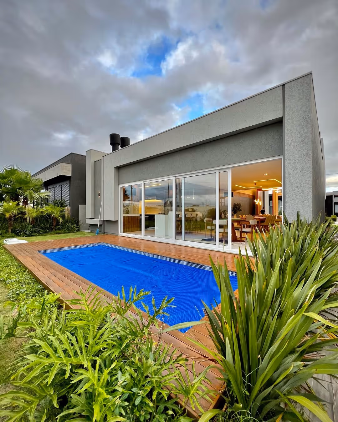 Casa moderna com piscina coberta por lona azul, deck de madeira, e jardim com plantas verdes ao redor.