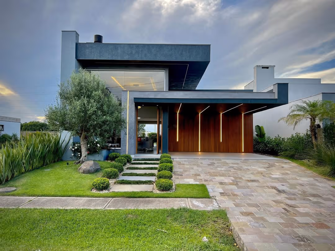 Casa moderna com fachada cinza escuro, garagem com iluminação embutida em ripas de madeira, jardim bem cuidado com arbustos, árvore e gramado verde.