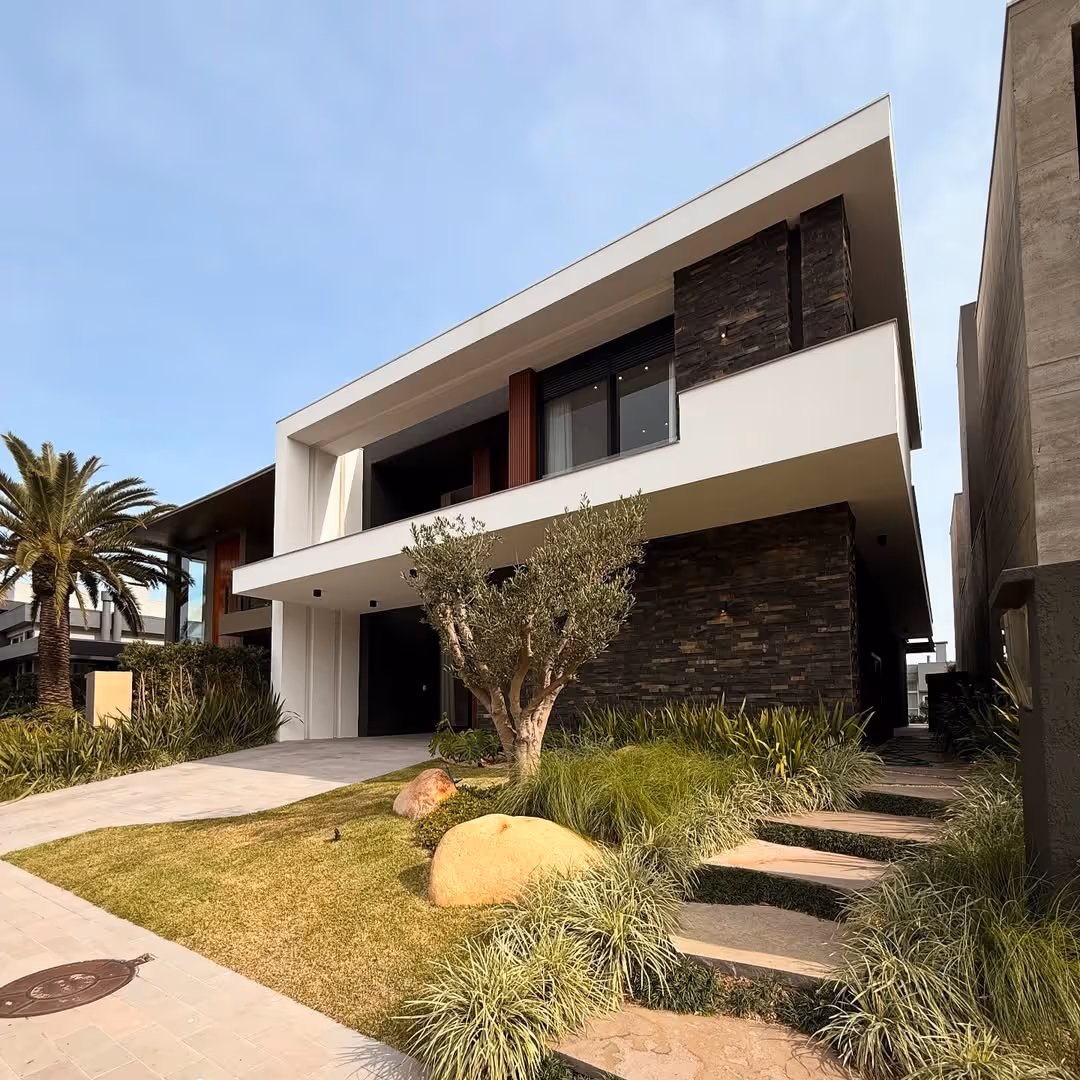 Casa moderna de dois andares com fachada branca e detalhes em pedra escura, jardim com grama, arbustos e árvores pequenas.