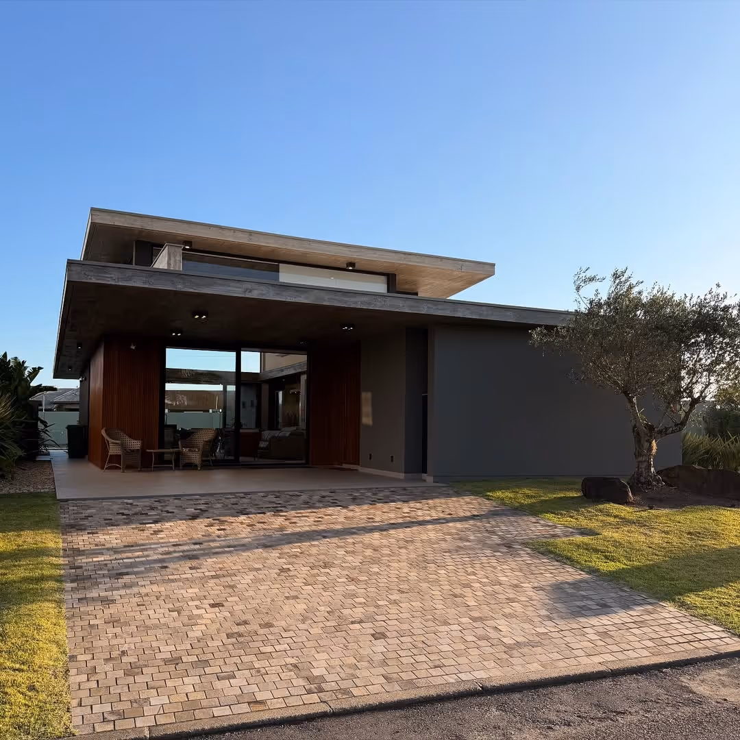 Casa moderna com telhado de concreto plano, varanda com duas cadeiras de vime, ampla entrada de calçada de pedra e árvore no jardim.