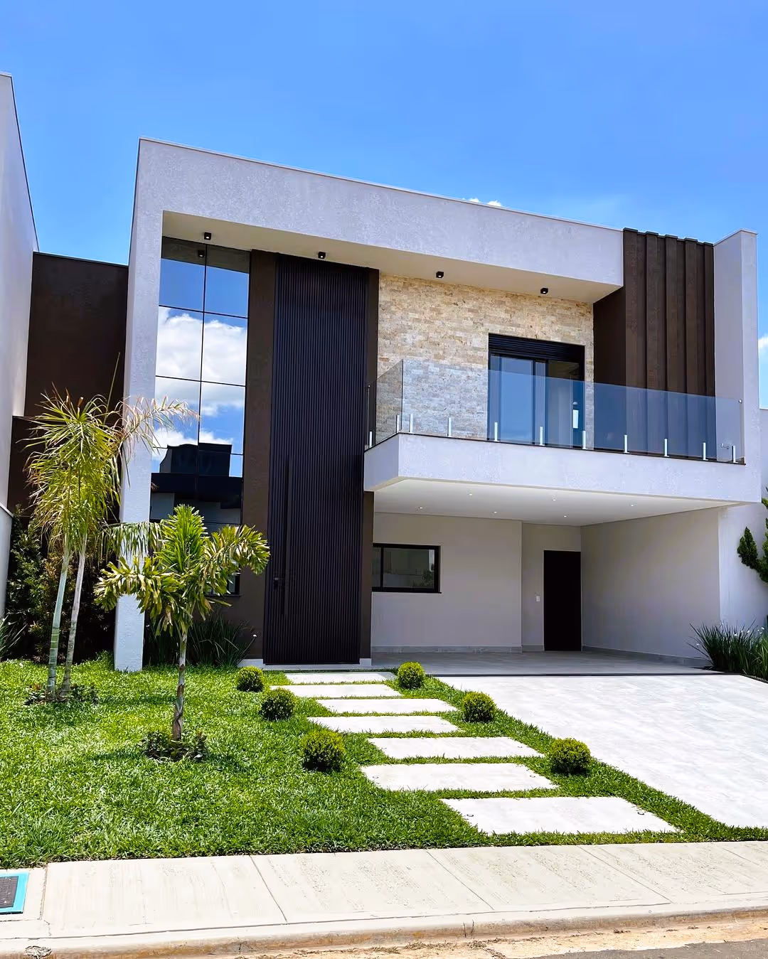 Casa moderna de dois andares com fachada clara, varanda com vidro, jardim gramado e palmeiras pequenas.