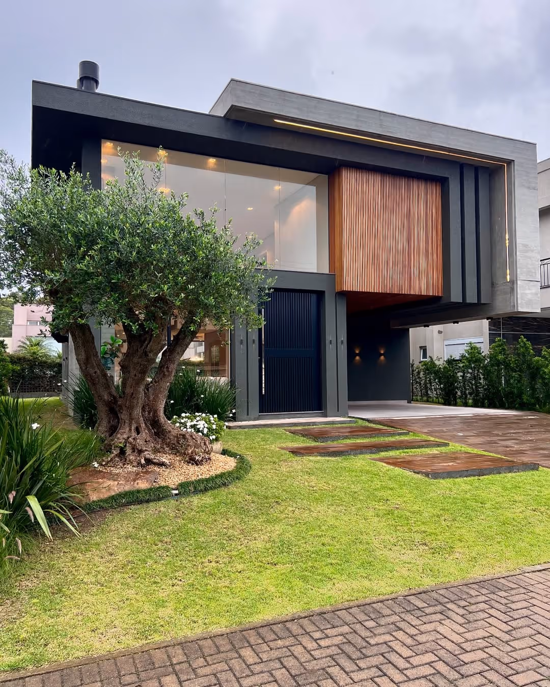 Fachada moderna de casa com grandes janelas de vidro, porta preta, detalhes em madeira e jardim com árvore e gramado bem cuidado.