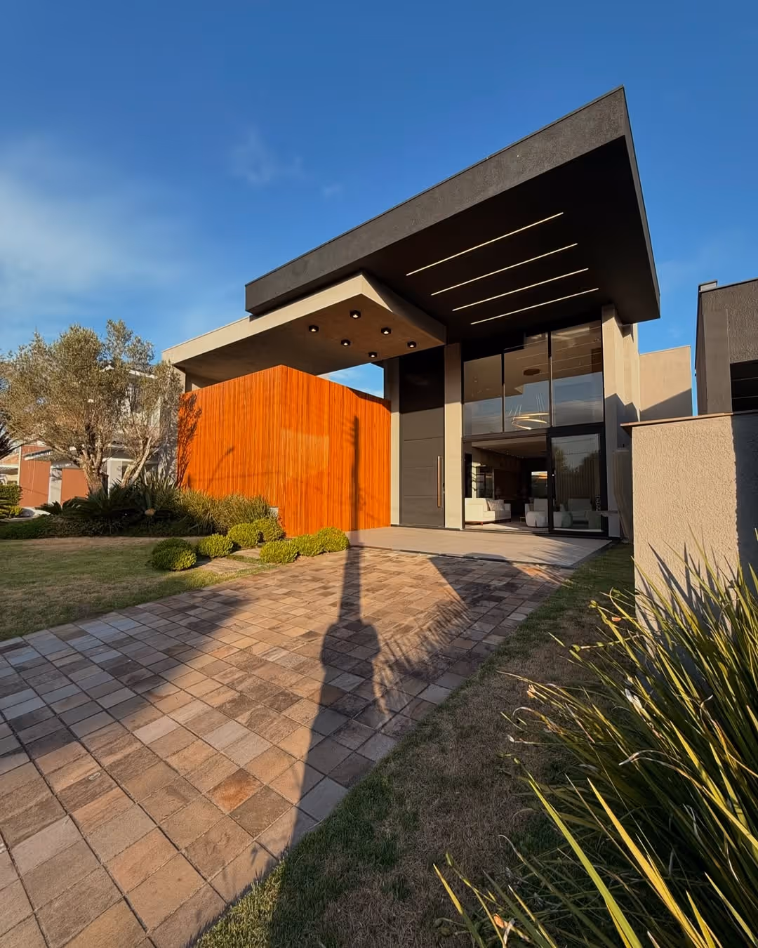 Fachada moderna de casa com parede de madeira, grande porta preta, teto saliente preto com iluminação embutida, e jardim ao redor.