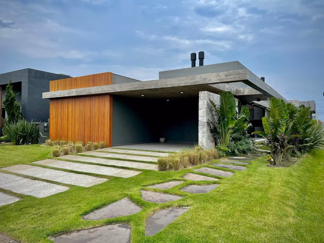 Casa moderna com fachada de concreto e madeira, garagem aberta e jardim com gramado verde e plantas tropicais.