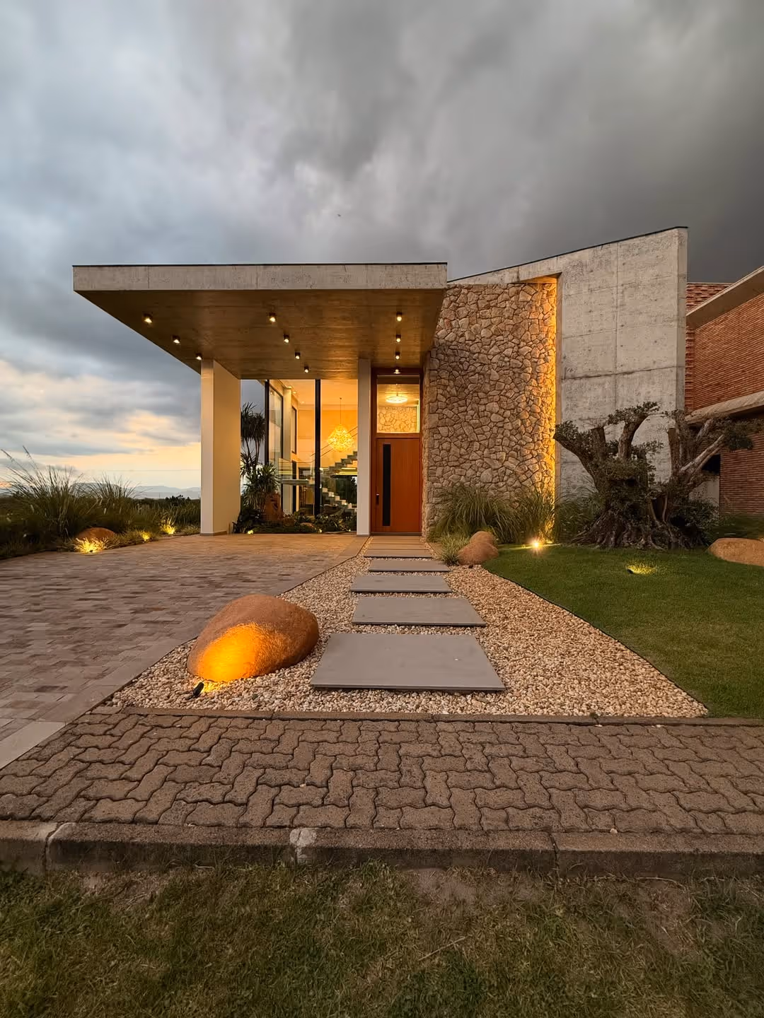 Casa moderna com entrada feita de pedras grandes iluminadas, caminho de placas cinzas, fachada com parede de pedras e concreto, e teto com luzes embutidas ao entardecer.