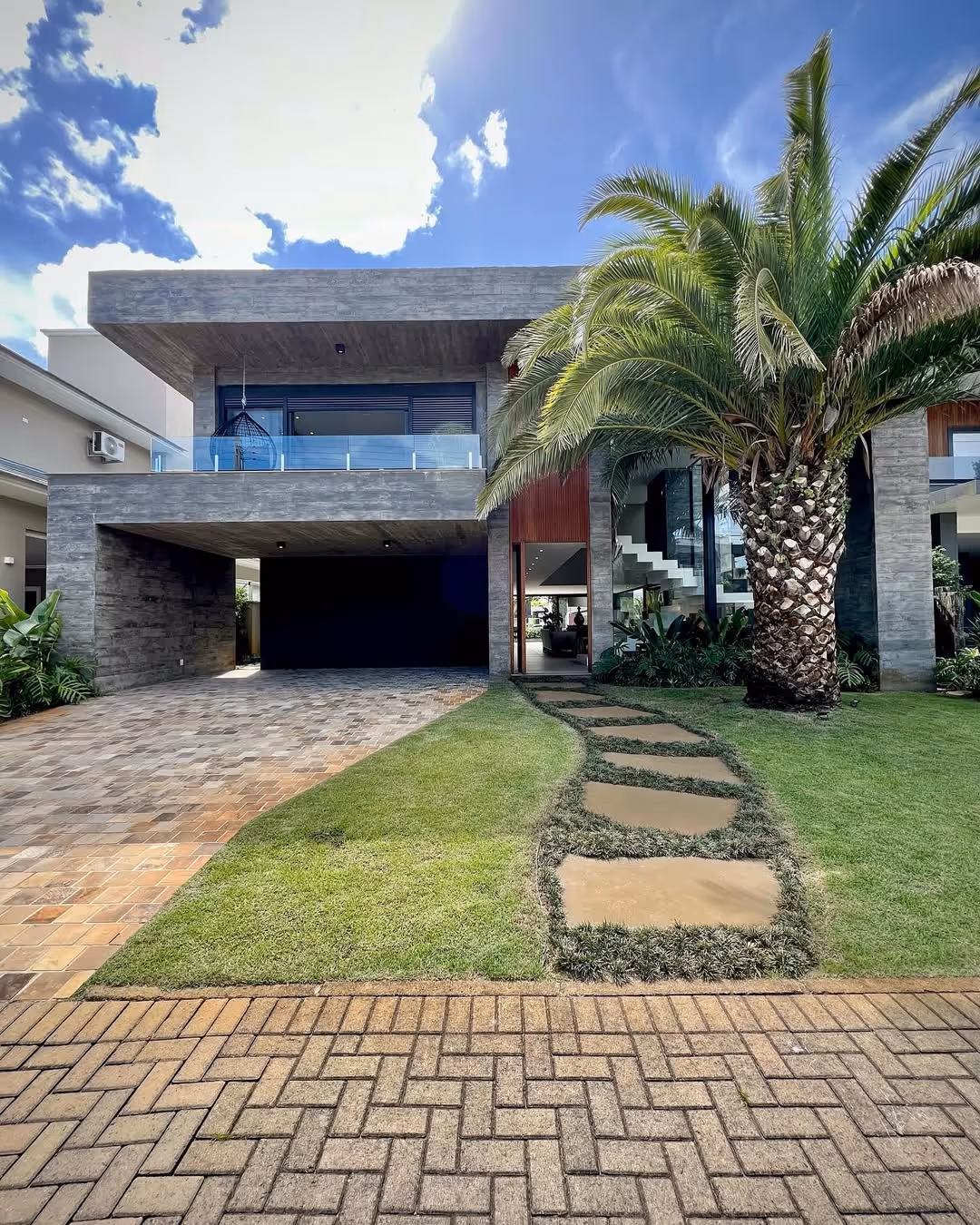 Casa moderna com estrutura de concreto, caminho de pedra em grama levando à entrada e uma grande palmeira no jardim frontal.