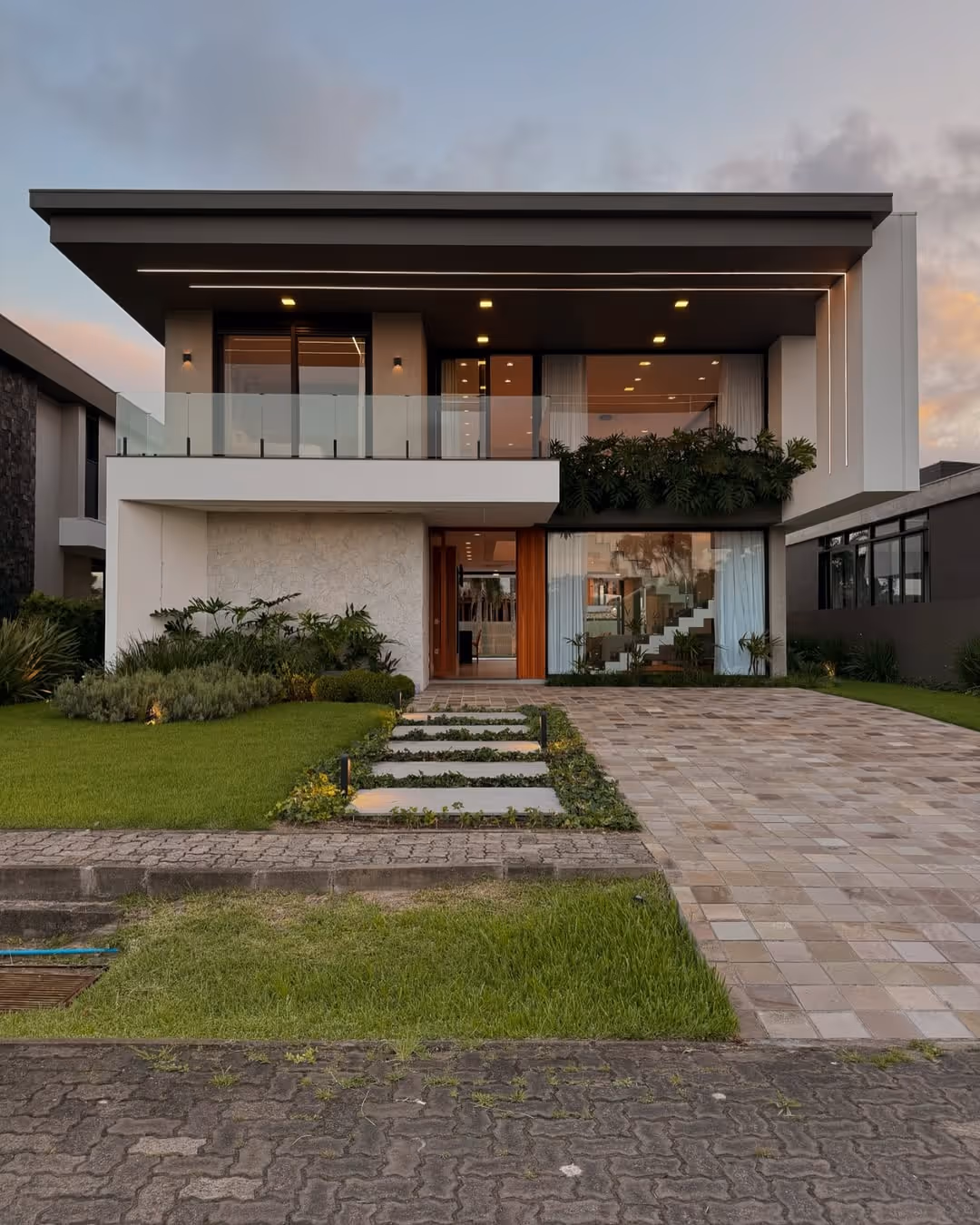 Fachada moderna de casa de dois andares com varanda de vidro, jardim frontal bem cuidado e grande vaga de garagem.