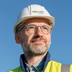 Homem de meia-idade com barba grisalha e óculos redondos sorrindo contra fundo cinza.