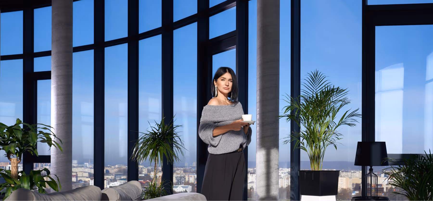 Mulher elegante segurando uma xícara de café em um ambiente moderno com grandes janelas de vidro e plantas internas.
