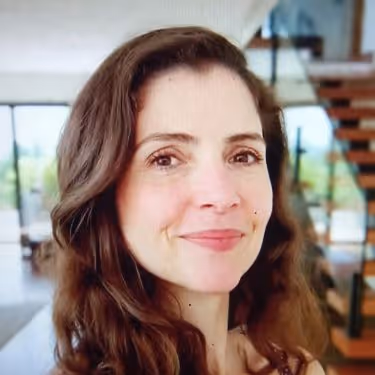 Mulher sorridente de cabelos castanhos ondulados em ambiente interno moderno com escada ao fundo.