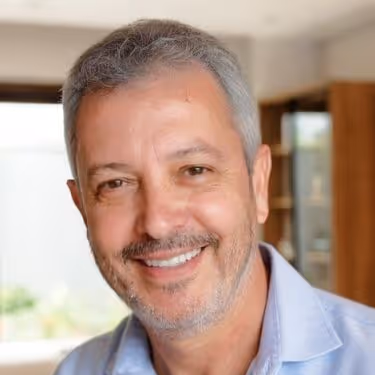 Homem sorridente de cabelos grisalhos com camisa azul clara em ambiente interno desfocado.