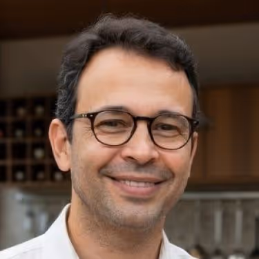 Homem de cabelos escuros curtos, usando óculos de armação preta, sorrindo em ambiente interno com prateleiras desfocadas ao fundo.